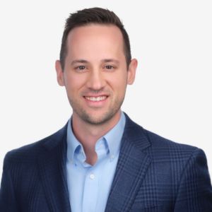 A smiling man with short dark hair, wearing a blue checkered blazer and light blue shirt, against a plain white background.