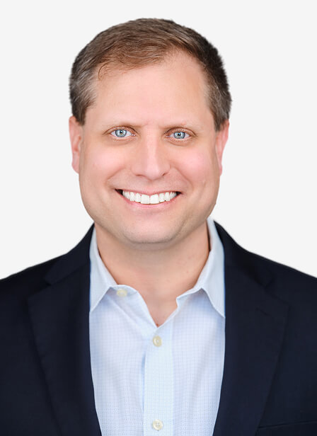 A smiling man with short light brown hair, blue eyes, wearing a dark blazer and light blue shirt against a plain white