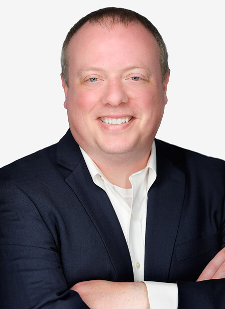 A smiling man in a dark suit with crossed arms, wearing a white shirt, against a plain light background.