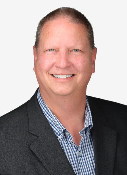 A smiling man in a dark suit and blue checkered shirt poses against a plain white background.