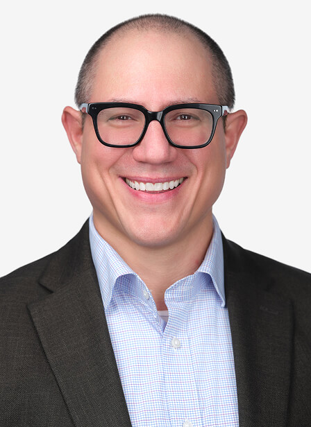 A smiling man with glasses, wearing a dark blazer and light blue shirt, against a plain white background.