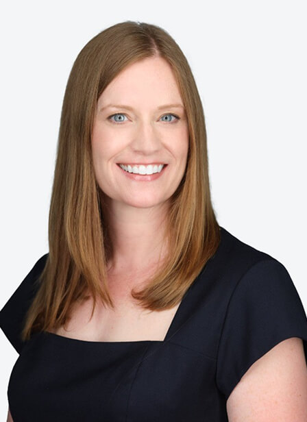 A woman with long, straight red hair, blue eyes, and a bright smile, wearing a dark navy top against a plain light