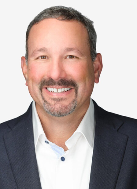 A smiling man with short dark hair, a beard, and mustache, wearing a dark suit jacket and white shirt against a light