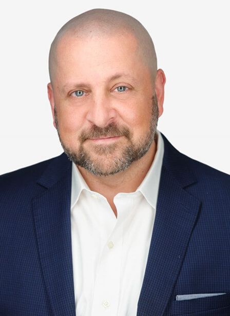 A bald man with a beard in a navy suit and white shirt, smiling confidently against a plain background.