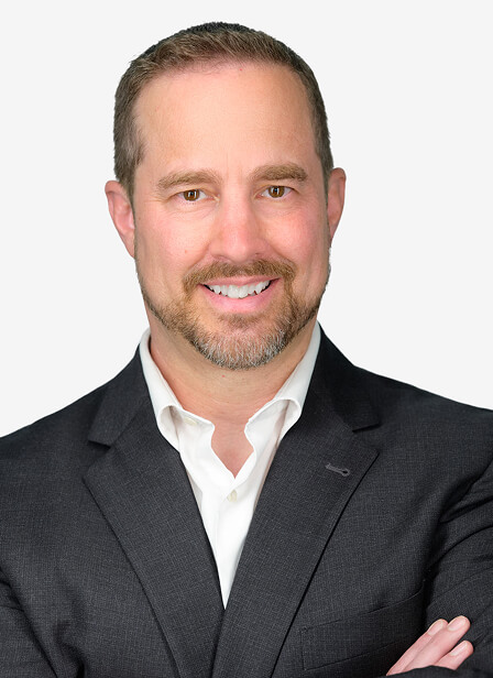 A professional man with short hair, a beard, wearing a dark suit and white shirt, smiling confidently against a light