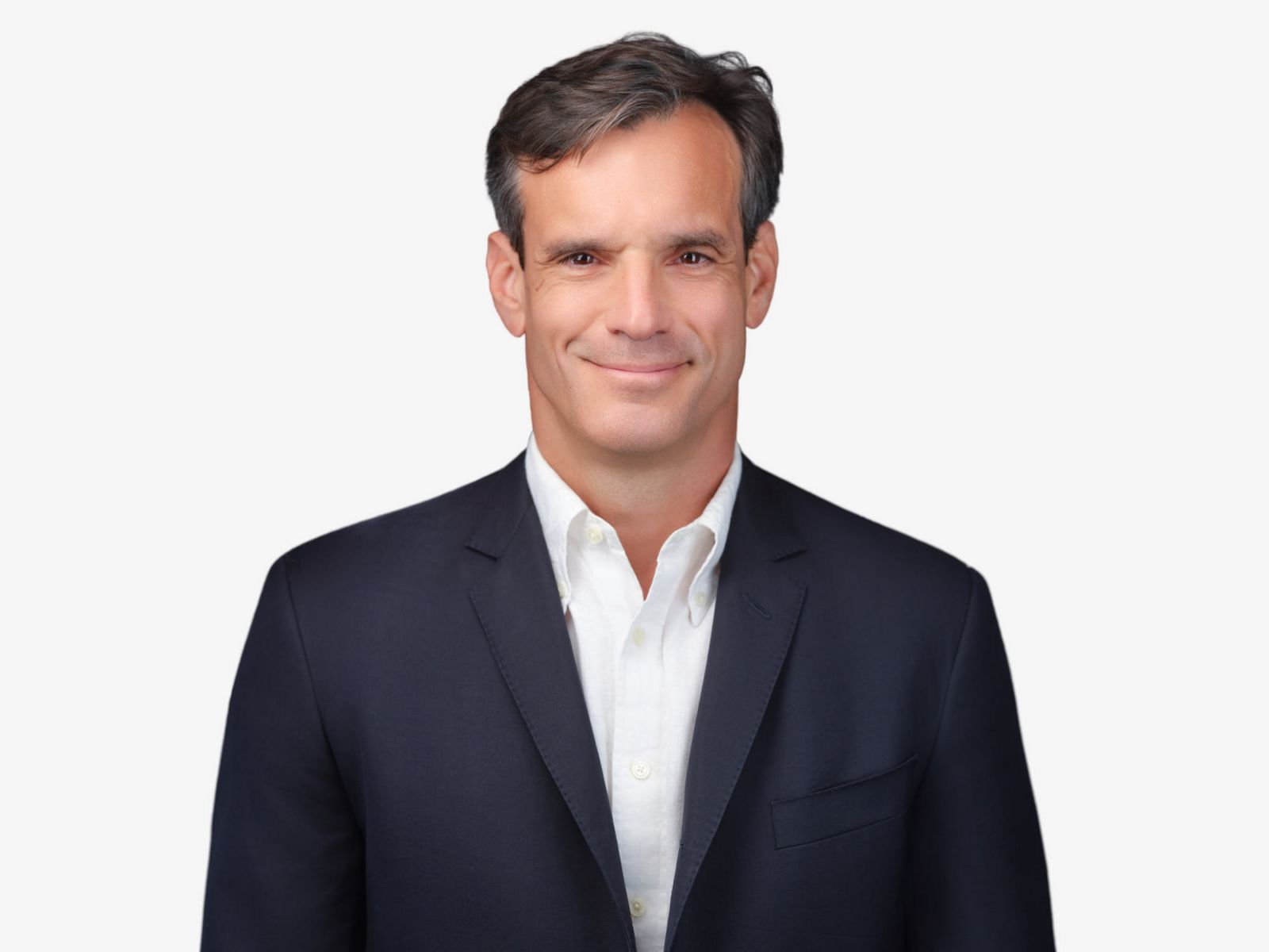 A man with dark hair, wearing a dark suit and white shirt, smiling against a plain light background.