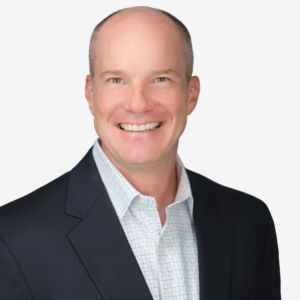 A smiling man in a dark suit and checkered shirt against a plain white background.