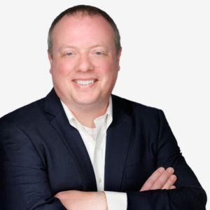 A smiling man in a dark suit with arms crossed, wearing a white shirt, against a plain white background.