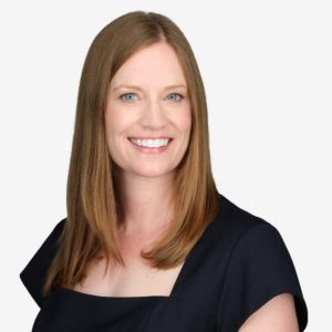 A woman with shoulder-length red hair, blue eyes, and a bright smile wearing a black top against a plain background.