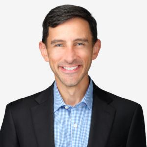 A smiling man with short dark hair, blue eyes, wearing a black blazer and blue checkered shirt, against a plain white